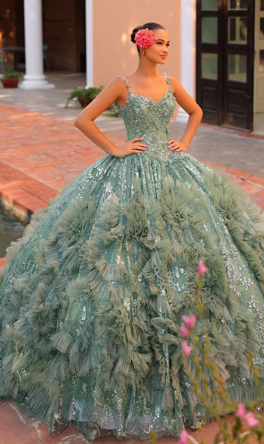 A woman stands outdoors wearing the Quinceanera Dress 54314 By Amarra, a detailed green v-neck ball gown with ruffled tulle and sequin embellishments, and has a pink flower in her hair.